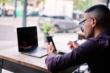 Seitenansicht eines gut aussehenden jungen hispanischen männlichen Unternehmers, der Informationen auf seinem Smartphone überprüft und Notizen in seinen Planer schreibt, während er an einem Tisch mit Laptop arbeitet - ADSF28568