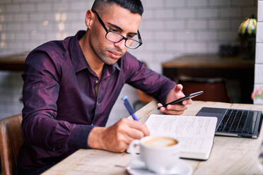Seitenansicht eines gut aussehenden jungen hispanischen männlichen Unternehmers, der Informationen auf seinem Smartphone überprüft und Notizen in seinen Planer schreibt, während er an einem Tisch mit Laptop arbeitet - ADSF28569