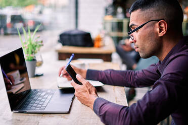 Seitenansicht eines gut aussehenden jungen hispanischen männlichen Unternehmers, der Informationen auf seinem Smartphone überprüft und Notizen in seinen Planer schreibt, während er an einem Tisch mit Laptop arbeitet - ADSF28570