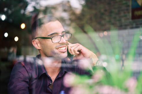 Kluger spanischer Geschäftsmann mit Brille, der sich Notizen auf einem Notizblock macht und einen Anruf entgegennimmt, während er in einer Cafeteria hinter dem Fenster sitzt - ADSF28571