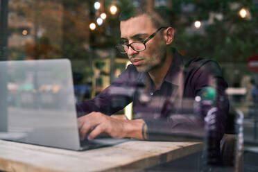 Seriöser hispanischer Manager in violettem Hemd und Brille tippt auf einer Netbook-Tastatur, während er an einem Tisch hinter dem Fenster sitzt und an einem Projekt in einem Café arbeitet - ADSF28572