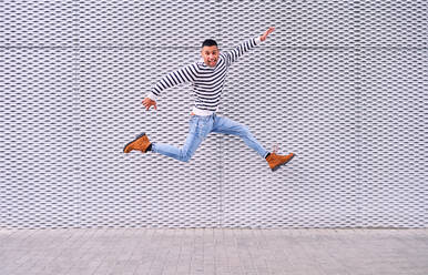 Full body excited Hispanic male smiling and leaping over pavement against ornamental gray wall while having fun on city street - ADSF28580