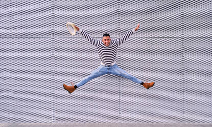 Full body excited Hispanic male smiling and leaping over pavement against ornamental gray wall while having fun on city street - ADSF28581