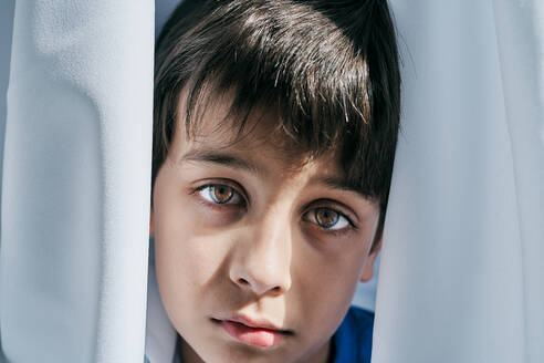 Unglücklicher kleiner Junge, der hinter einem Vorhang hervorschaut, während er unter häuslicher Gewalt leidet und sich vor seinen Eltern versteckt - ADSF28584