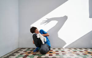 Scared little boy with toy in hands sitting near wall with shadow of angry violent parent with raised arm - ADSF28588