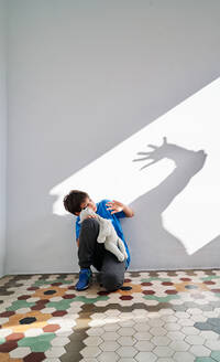 Verängstigter kleiner Junge mit Spielzeug in den Händen sitzt in der Nähe der Wand mit Schatten der wütenden gewalttätigen Eltern mit erhobenem Arm - ADSF28589