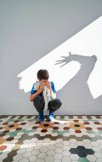Verängstigter kleiner Junge mit Spielzeug in den Händen sitzt in der Nähe der Wand mit Schatten der wütenden gewalttätigen Eltern mit erhobenem Arm - ADSF28590