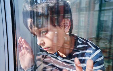 Through glass of unhappy preteen boy with bruises on face looking away while standing near window at home as concept of domestic violence and child abuse - ADSF28597