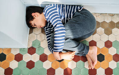 From above of unhappy lonely preteen boy lying on floor and embracing knees as victim of domestic violence and abuse - ADSF28598