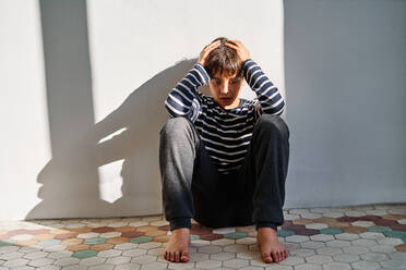 Unhappy kid sitting on floor and covering head with hands while suffering from domestic violence at home - ADSF28600
