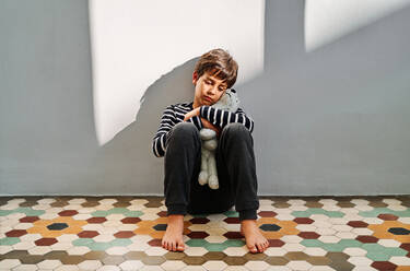 Upset lonely preteen boy victim of domestic violence sitting on floor and embracing toy - ADSF28602