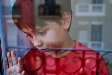 Durch Glas von unglücklichem Jungen mit blauen Flecken im Gesicht, der wegschaut, während er zu Hause am Fenster steht, als Konzept von häuslicher Gewalt und Kindesmissbrauch - ADSF28608