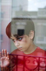 Durch Glas von unglücklichem Jungen mit blauen Flecken im Gesicht, der wegschaut, während er zu Hause am Fenster steht, als Konzept von häuslicher Gewalt und Kindesmissbrauch - ADSF28609