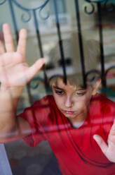 Through glass of unhappy preteen boy with bruises on face looking away while standing near window at home as concept of domestic violence and child abuse - ADSF28610