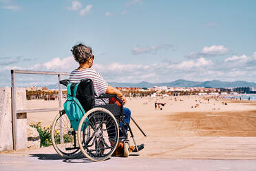 Side view of handicapped female in protective mask sitting in wheelchair with backpack and enjoying sunny summer day on sandy beach - ADSF28615