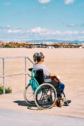 Side view of handicapped female in protective mask sitting in wheelchair with backpack and enjoying sunny summer day on sandy beach - ADSF28616