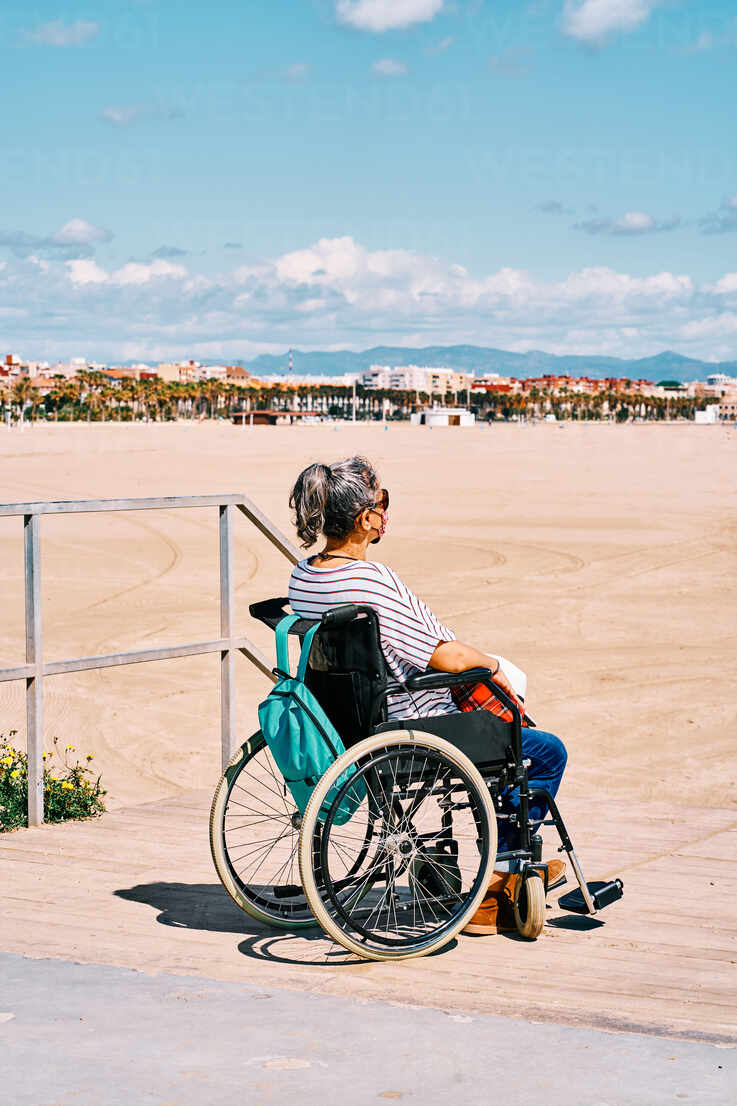 Seitenansicht einer behinderten Frau mit Schutzmaske, die mit Rucksack im  Rollstuhl sitzt und einen sonnigen Sommertag