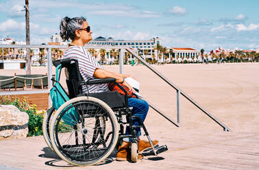 Side view female traveler in wheelchair with backpack enjoying summer journey on beach near blue sea - ADSF28617