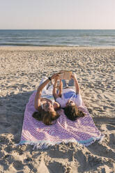 Junge Freundinnen lesen ein Buch und liegen auf einer Picknickdecke am Strand - JRVF01597