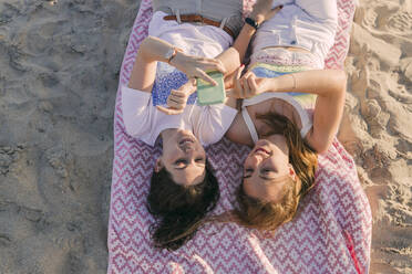 Frauen benutzen ein Mobiltelefon, während sie auf einer Picknickdecke am Strand liegen - JRVF01600