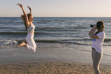 Frau fotografiert ihre Freundin beim Springen am Strand - JRVF01603