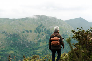 Rückenansicht einer anonymen älteren Frau mit Rucksack und Wanderstock, die auf einem grasbewachsenen Hang in Richtung Berggipfel spazieren geht, während eines Ausflugs in die Natur - ADSF28627