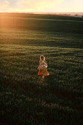 Young female in vintage rustic style dress running alone in vast green grassy field in summer evening in countryside - ADSF28629