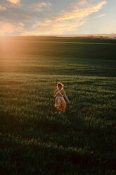 Young female in vintage rustic style dress running alone in vast green grassy field in summer evening in countryside - ADSF28630