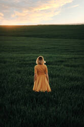 Junge Frau im Vintage-Stil, die nachdenklich wegschaut, während sie allein auf einem grasbewachsenen Feld bei Sonnenuntergang im Sommerabend auf dem Land steht - ADSF28631