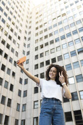 Junge Frau macht Friedenszeichen, während sie ein Selfie vor einem Gebäude macht - MTBF01087