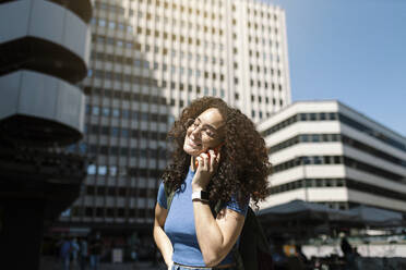 Smiling woman in eyeglasses talking on smart phone near building - MTBF01089