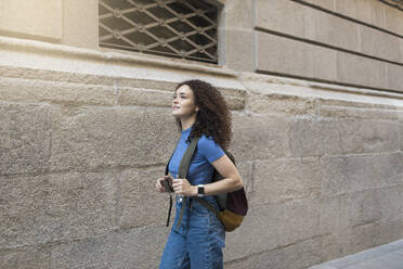 Young woman with backpack walking by wall - MTBF01093