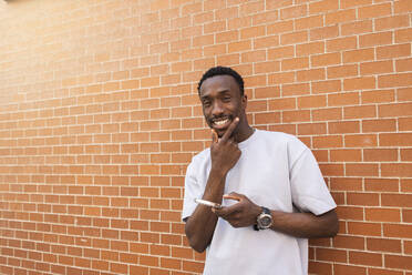 Junger Mann hält Smartphone und lächelt vor einer Backsteinmauer - MTBF01103