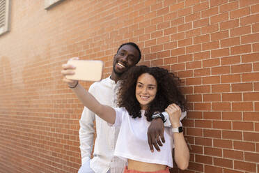 Young man and woman taking selfie through smart phone in front of brick wall - MTBF01107