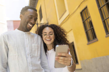 Lächelnder Mann schaut auf ein Smartphone, das von einer Frau in der Nähe eines Gebäudes gehalten wird - MTBF01122