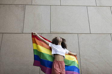 Woman holding rainbow flag while standing in front of wall - MTBF01133