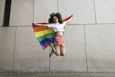 Cheerful woman jumping while holding rainbow flag in front of wall - MTBF01134