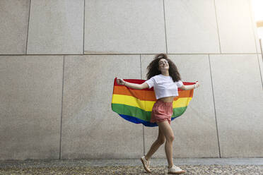 Woman holding rainbow flag while walking in front of wall - MTBF01135