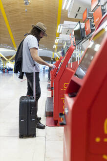 Junger Mann mit Bart, langen Haaren und Hut beim Kauf von Flugtickets am Automatenterminal der Fluggesellschaft - ADSF28747