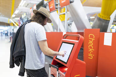 Junger Mann mit Bart, langen Haaren und Hut beim Kauf von Flugtickets am Automatenterminal der Fluggesellschaft - ADSF28748