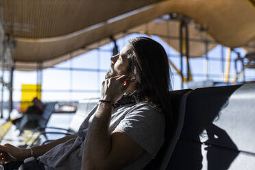 Der Typ mit dem Hut am Flughafen, der im Warteraum sitzt und auf seinen Flug wartet, setzt kabellose Kopfhörer auf, um Musik zu hören - ADSF28750