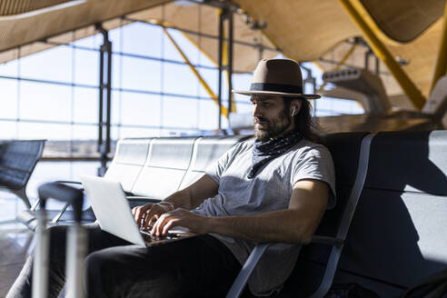 Der Typ mit dem Hut am Flughafen im Warteraum, der auf seinen Flug wartet, mit kabellosen Kopfhörern, um Musik zu hören, während er mit seinem Laptop arbeitet - ADSF28751