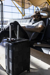 Der Typ mit Hut am Flughafen im Wartesaal, der auf seinen Flug wartet, mit kabellosen Kopfhörern Musik hört, schläft und mit seinem Hut die Augen verdeckt - ADSF28753