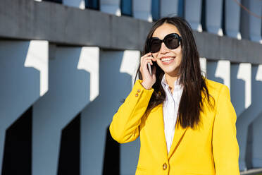 Lächelnde asiatische Geschäftsfrau mit gelbem Mantel und Smartphone zu Fuß auf der Straße mit Gebäude im Hintergrund - ADSF28754