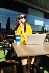 Smiling asian business woman with yellow coat sitting at a table having coffee with her smart phone and laptop - ADSF28759