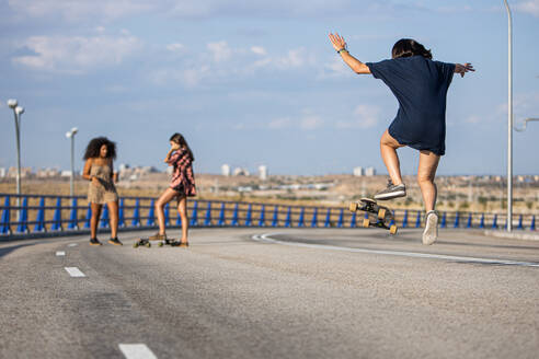 Unbekannte junge Frau, die an einer Brücke einen Trick mit ihrem Longboard macht, mit ihren Begleitern im Hintergrund - ADSF28769
