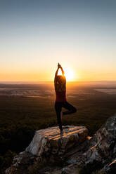 Young yogi woman practicing yoga on a rock in the mountain with the light of sunrise, back view with one leg and arms lifted - ADSF28798