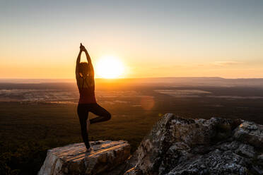 Young yogi woman practicing yoga on a rock in the mountain with the light of sunrise, back view with one leg and arms lifted - ADSF28799