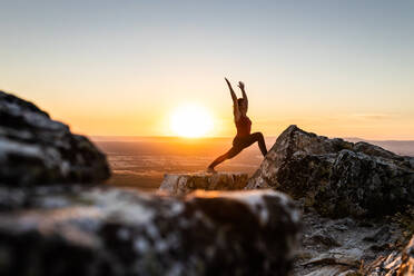 Junge Yogi Frau übt Yoga auf einem Felsen in den Bergen mit dem Licht des Sonnenaufgangs, Seitenansicht mit einem Bein auf einem Felsen und Arme erhoben - ADSF28800