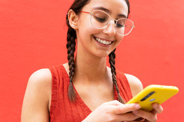 Fröhliche junge Frau in Zöpfen Frisur Surfen auf Smartphone stehend auf rotem Hintergrund in der Straße - ADSF28804
